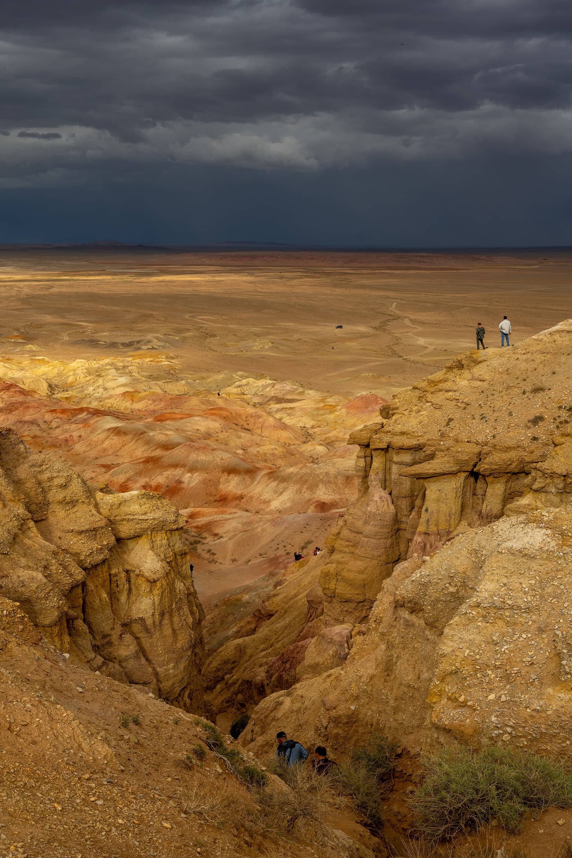 Layered ochre and red canyon walls beneath a dark storm sky, with two small figures on the rim