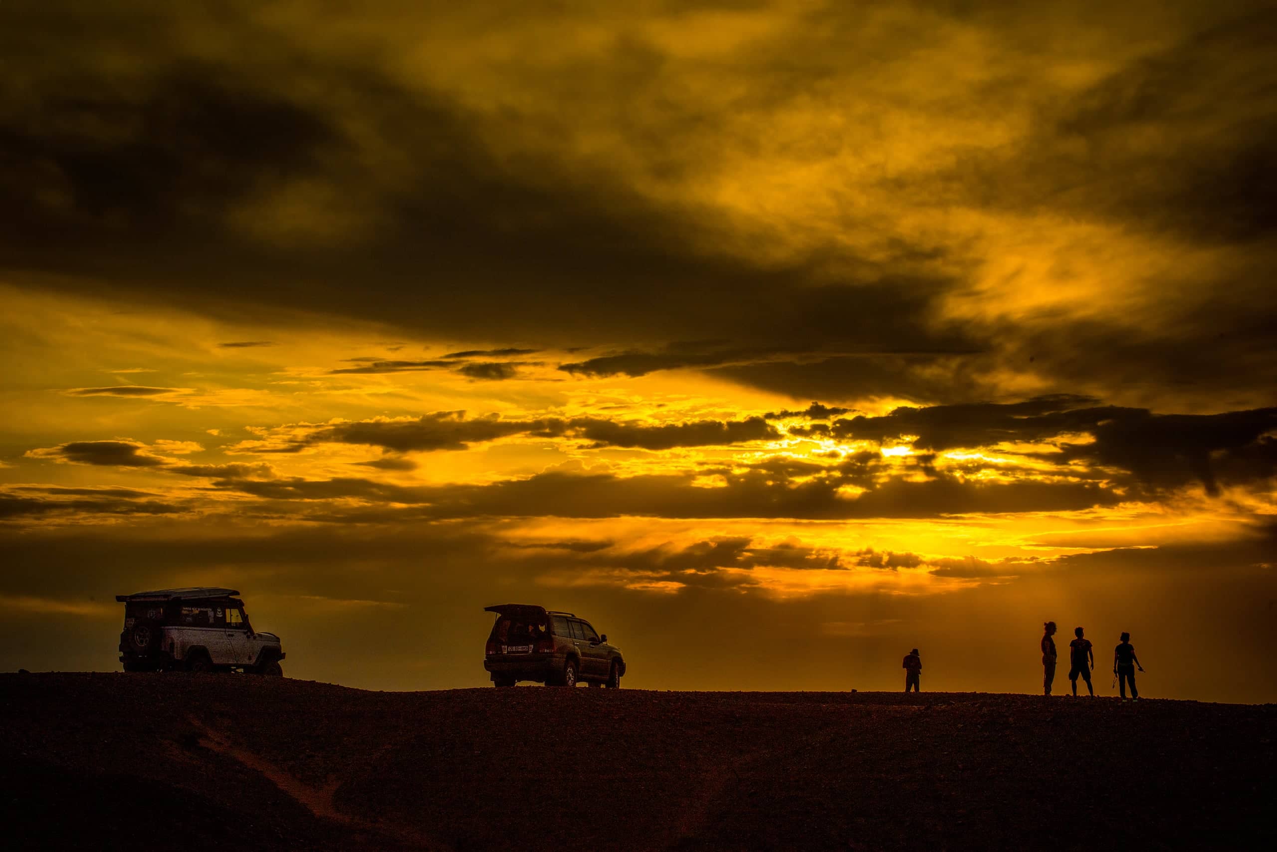 Expedition vehicles and travellers silhouetted on a Gobi ridge under a burning orange sunset sky