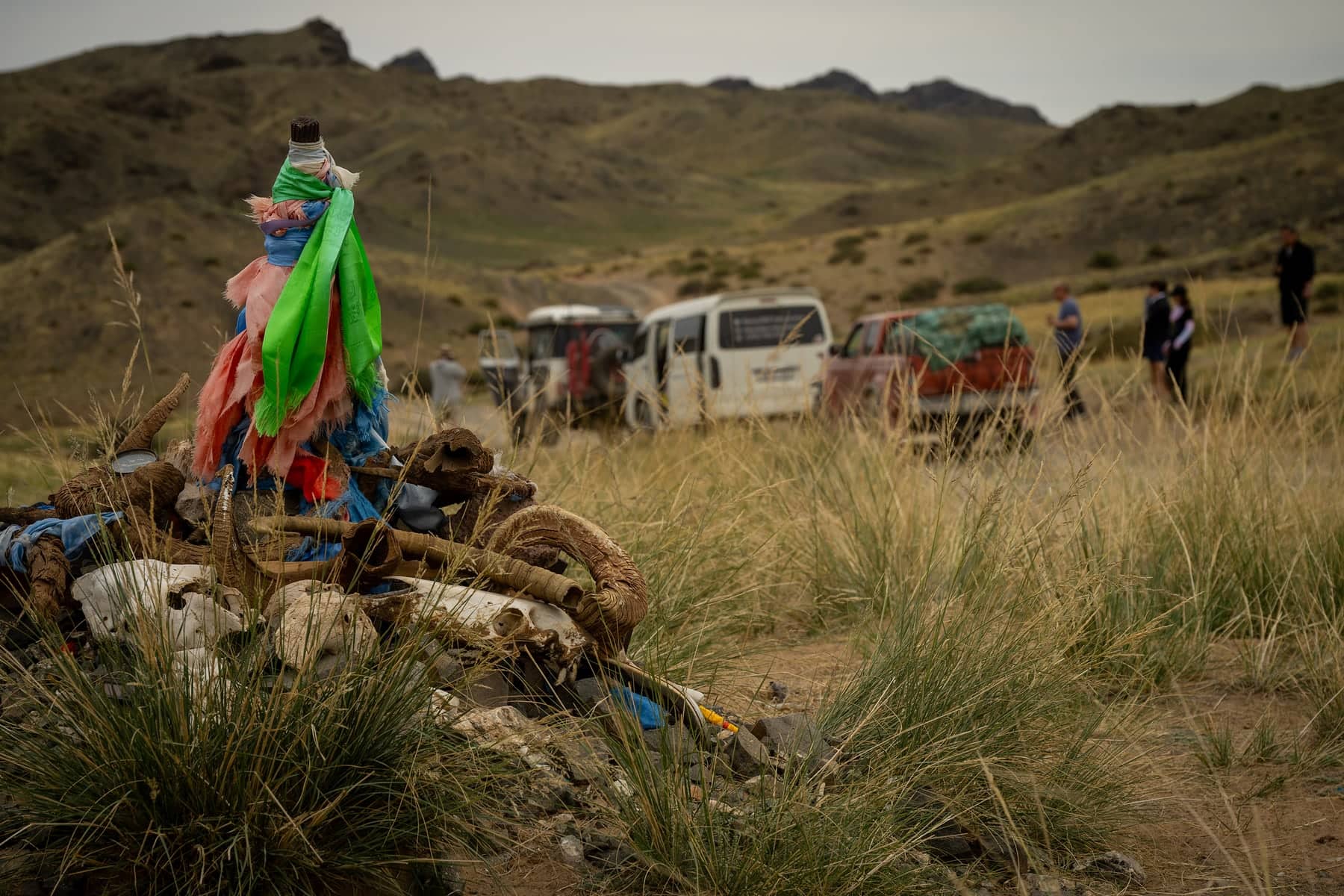 A roadside ovoo cairn draped in blue and green ceremonial scarves, with expedition vehicles in the grassland valley behind