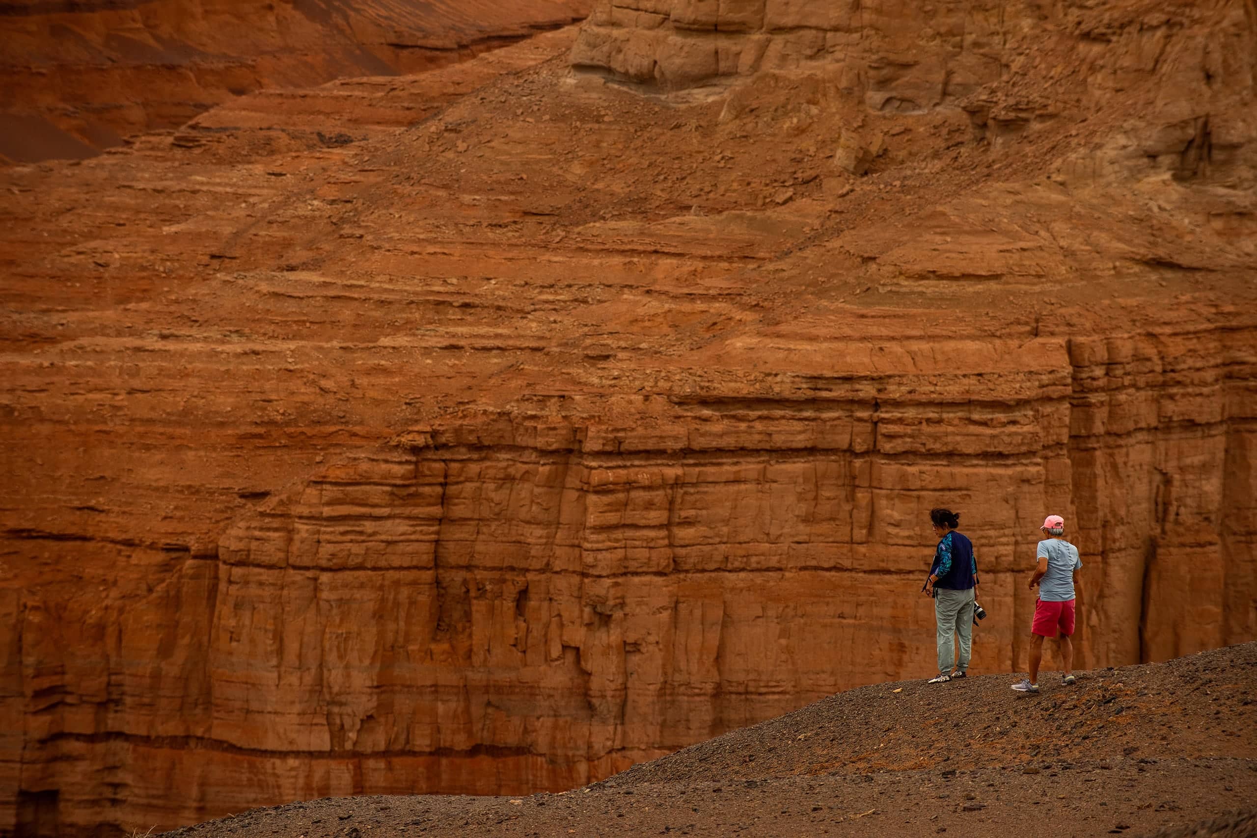 Two travellers dwarfed by layered red canyon cliffs in the Gobi