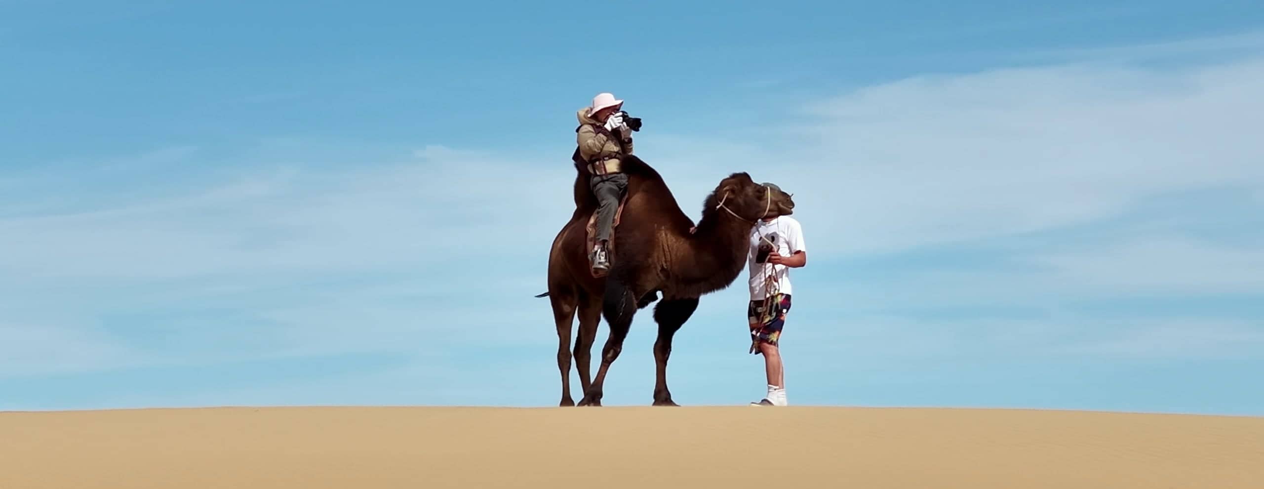 A photographer on a Bactrian camel raises a camera on a dune crest while a guide stands beside the camel