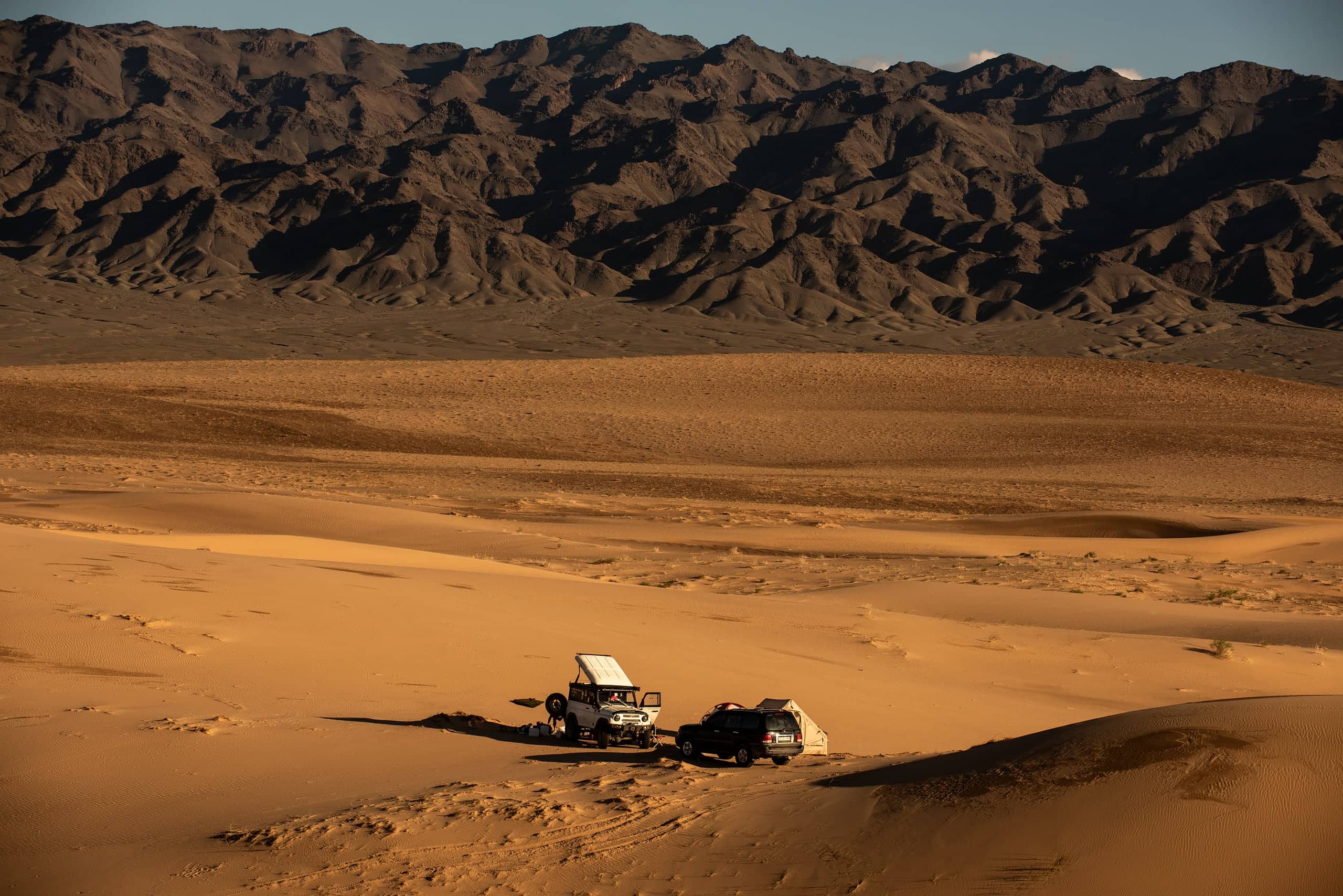 Two expedition vehicles parked on golden Gobi sand below a dark, rugged mountain range